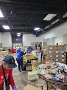 Volunteers sort and pack food donations at tables inside a large warehouse, surrounded by stacked boxes and packaged bread.